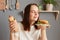 Portrait of joyful satisfied woman with dark hair dressed in white T-shirt holding big burger sandwich and sausage in dough,