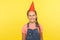 Portrait of joyous adorable little girl in denim overalls with funny cone on head smiling at camera, enjoying birthday party