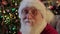 Portrait kind smiling Santa Claus with gray beard in suit and gold glasses sitting in chair on background of Christmas