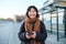 Portrait of Korean woman in winter jacket, standing with smartphone, waiting for bus on stop, looking at mobile phone