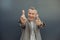 Portrait of laughing aged gray haired businessman standing in stylish suit and pointing fingers forward on gray