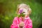 Portrait of little 3 years old blonde girl in pink jacket hiding behind branch of purple lilac in hand. Lilac and unrecognizable