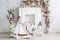 Portrait of a little blonde girl in flowers. She stand near the wooden horse rocker in the white flowers room