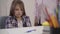 Portrait of little caucasian schoolgirl sitting at the table with upset facial expression as her mother shaking pen in