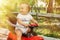 Portrait of a little cheerful baby in white shirt sitting half turned back on red push car in park or garden in sun rays