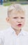 Portrait of a little displeased and dissatisfied boy with golden blonde hair in sunny summer day on green blurred