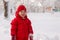 Portrait of a little girl 3 years old in mittens, a red jacket and a knitted hat, who stands in a snowy park in winter