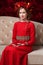 Portrait of a little girl in a flower wreath and red dress sitting on the couch