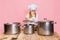 Portrait of little girl in white cook uniform and huge chef& x27;s hat at kids kitchen with big pans isolated on pink studio