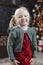 Portrait of little jolly blonde girl in festive new year clothes, traditional xmas decorated living room. Vertical frame