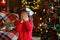 Portrait of a little surprised girl in a festively decorated room with lights of garlands for Christmas. A child in red pajamas