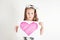 Portrait of little thoughtful girl with long hair holding in hands sheet of paper with pink heart on white background.