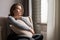 Portrait of lonely sad Ñaucasian young woman sitting near window and hugs pillow.