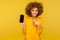 Portrait of lovely curly-haired woman with engaging smile, wearing urban style hoodie, pointing mobile phone