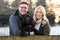 Portrait Of Loving Couple With Pet Spaniel Dog Leaning On Fence On Snowy Walk In Winter Countryside