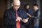 Portrait of male with glasses bald businessman using phone in hand, dressed striped suit and red tie, stands indoors