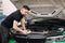 Portrait of a male mechanic working in his garage. Young bearded man checks the engine in the car, the hood is open