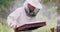 Portrait male, young, bearded, beekeeper in white protective suit puts a smoking smoker on the hive aside and which is