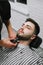 Portrait of a man in barber shop, lying in a chair and looking up while the barber cuts his beard. Barber trims his beard