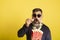 Portrait of man with white beard and sunglasses eating popcorn astonished watching television on yellow background