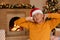 Portrait of mature man before christmas, sitting near fireplace and xmas tree and stretching hands, looks sleepy, wearing casual