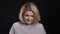 Portrait of middle-aged blonde short-haired overweight business woman watching shyly into camera on black background.