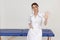 Portrait of middle-aged confident masseuse woman standing near blue couch, stretching hand forward with open palm.