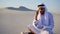 Portrait of Muslim designer Arabian Sheikh who coordinates on mobile with client building and sits on sand with project