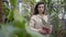 Portrait of mysterious Caucasian woman standing between green plants holding pot with cactus. Elegant brunette brown