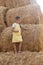 Portrait of nice young female kid standing on haystack thinking eyes looking down and holding hay in one hand wearing