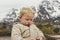 Portrait of nordic blond boy sitting on the floor warm with a coat playing outdoors with snowy mountains behind