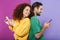 Portrait of optimistic caucasian couple in colorful clothing smiling and holding cellphones standing back to back