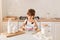 Portrait of optimistic little cute female kid with dark hair and pigtails wearing white t shirt cooking in light kitchen, mixing