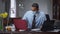 Portrait of overworking African American man in Covid face mask typing on laptop and tablet keyboards, holding head in