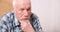 Portrait of a pensive elderly man with gray hair touching his beard and looking to the side sitting on the couch at home.