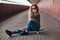 Portrait of a pensive girl in sunglasses dressed in a hoodie and ripped jeans sitting on a skateboard at bridge footway.