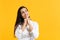 Portrait of pensive smiling young woman in white shirt put hand prop up on chin, looking up  on bright yellow