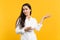 Portrait of perplexed young woman in white casual shirt looking camera, pointing hands aside isolated on bright yellow