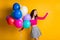 Portrait photo of happy female student holding air balloons smiling keeping hand up isolated on bright yellow color