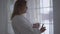 Portrait of plump girl in white bathrobe standing near large floor-to-ceiling window with cup of coffee or tea. Morning