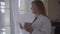 Portrait of plus size girl in white bathrobe standing near large floor-to-ceiling window with cup of coffee or tea