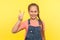 Portrait of positive adorable little girl with braid in denim overalls showing v sign with double fingers