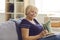 Portrait of positive aged woman granny in t-shirt sitting on sofa with glasses and book