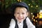 Portrait of positive little boy in cap against backdrop of Christmas tree with garland lights. Sincere child emotions. Happy child