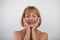 Portrait of positive lovely senior lady smiling, closing her eyes and touching face over light studio background