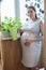 Portrait of pregnant gardener with watering can stands on domestic balcony with potted flowers, one woman