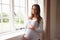 Portrait Of Pregnant Woman Standing By Window Holding Belly