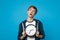 Portrait of preoccupied young man in casual clothes holding round clock isolated on blue wall background in studio. Time