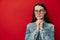 Portrait of pretty calm young woman in eyewear keeps hands together near face, looks optimistic aside, dressed in denim jacket