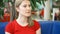 Portrait of pretty dreamy young woman in red t-shirt sitting in cafe thinking. Blue sofa green tree